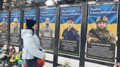 Оксана Шелудько біля стенду на Алеї Слави у Новій Водолазі, на якому розміщене фото її чоловіка Оксана Шелудько біля стенду на Алеї Слави у Новій Водолазі, на якому розміщене фото її чоловіка