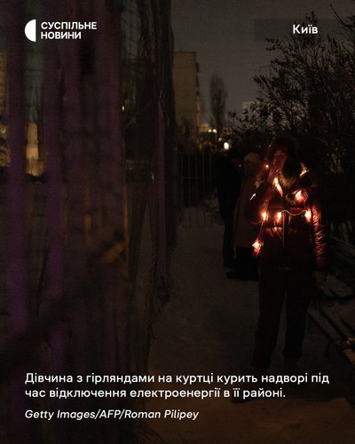 Дівчина з гірляндами на куртці курить надворі під час відключення електроенергії в її житловому районі у Києві Дівчина з гірляндами на куртці курить надворі під час відключення електроенергії в її житловому районі у Києві