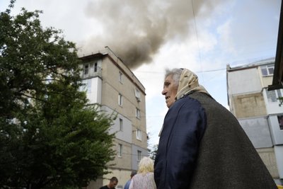 Літня жінка стоїть на фоні багатоповерхівки, в яку влучив російський реактивний безпілотник Літня жінка стоїть на фоні багатоповерхівки, в яку влучив російський реактивний безпілотник