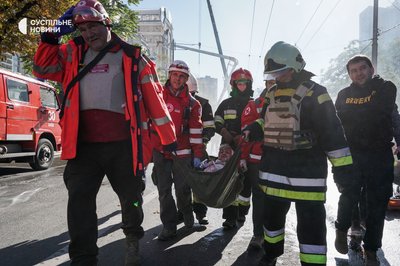 Рятувальники несуть врятовану з-під завалів будинку 84-річну жінку, Київ, жовтень 2022 року. В руках вона тримає тканинний вузлик з уцілілими речами.
